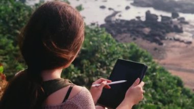 Genç Asyalı kadın deniz kenarındaki tepede otururken resim çizmek için dijital tablet kullanıyor.