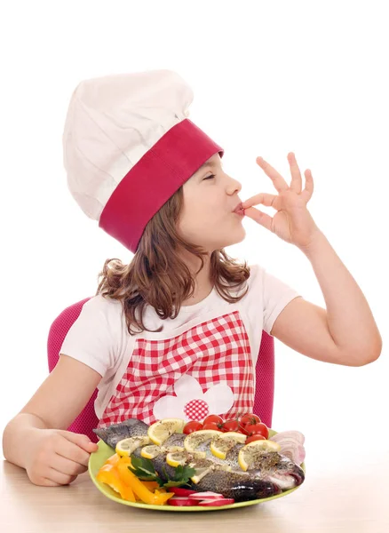 Счастливая маленькая девочка готовит с рыбой-тройкой и ласковым знаком руки — стоковое фото