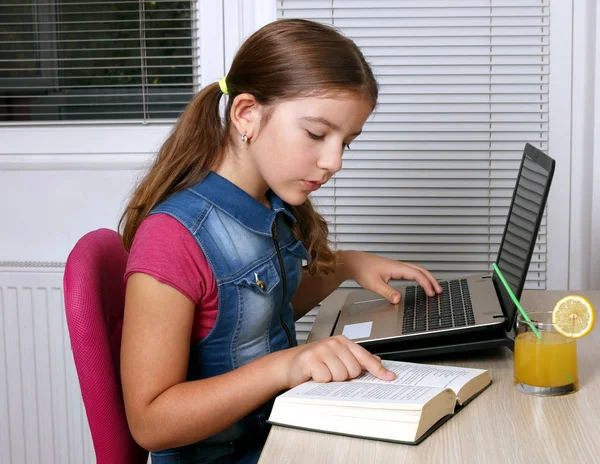 有一本书和一台手提电脑在家里的小女孩学习。 — 图库照片
