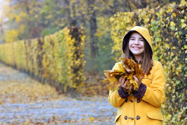 Fröhliches Mädchen im gelben Kapuzenmantel hält die Blätter in ihren Händen — Stockfoto