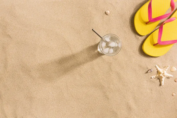 Summer fashion, summer outfit on sand background. Yellow flip flops, glass of water and seashells. Flat lay, top view. Harsh light with shadows