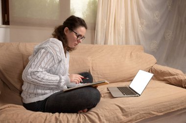 Oturma odasındaki kanepede oturan ve evde uzaktan çalışan genç bir bayan. Dizüstü bilgisayarı ve kitaplarıyla çalışıyor.