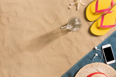 Yaz tatili kompozisyonu. Parmak arası terlikler, şapka, akıllı telefon ve kumlu zeminde bir bardak su. Seyahat tatili konsepti. Yaz dönemi. Havludan yapılmış kenar bileşimi