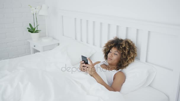 Улыбающаяся женщина расслабляется с телефоном в постели — стоковое видео