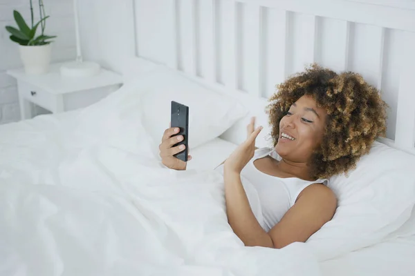 Mujer sonriente en la cama hablando en línea —  Fotos de Stock