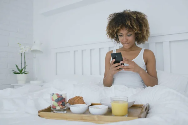 Mulher com smartphone na cama tendo refeição — Fotografia de Stock