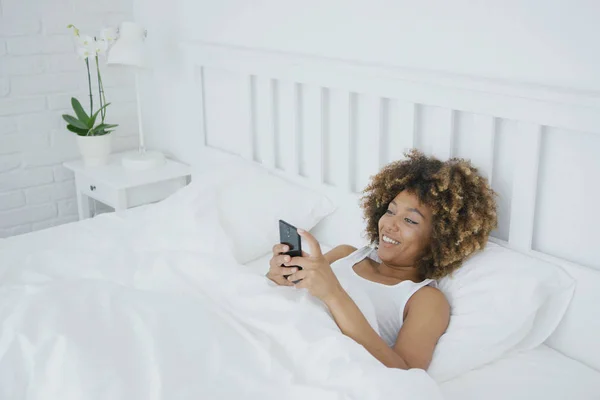 Lächelnde Frau entspannt sich mit Handy im Bett — Stockfoto