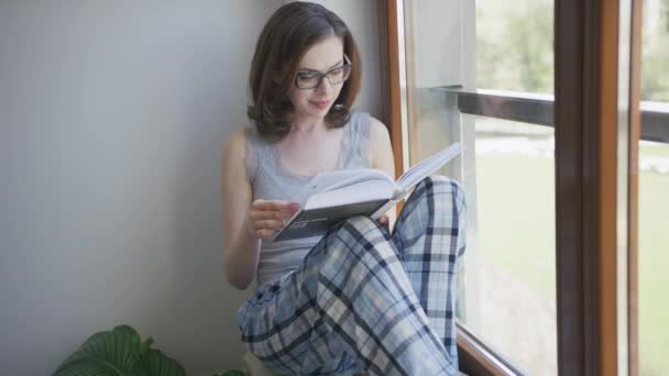 Młoda kobieta siedzi w okna w domu i czytanie — Wideo stockowe