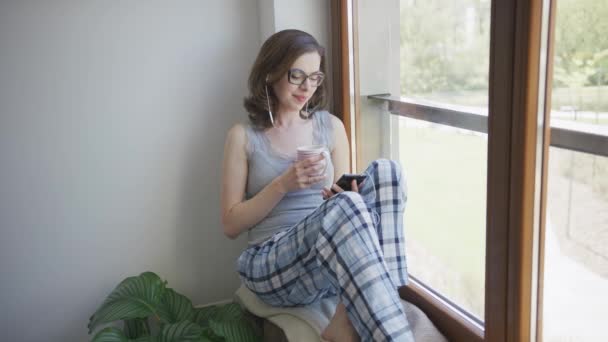 Улыбающаяся женщина сидит в окне, используя смартфон и наушники — стоковое видео