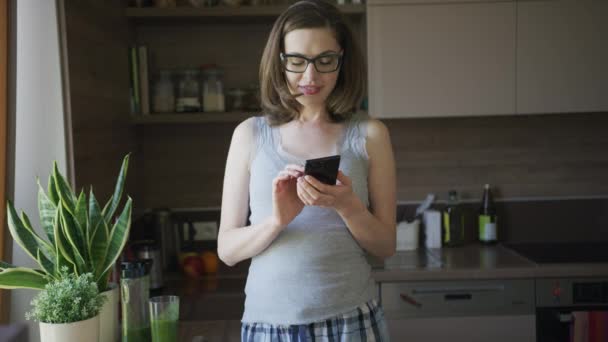 Aantrekkelijke vrouw met behulp van smartphone staande in keuken — Stockvideo