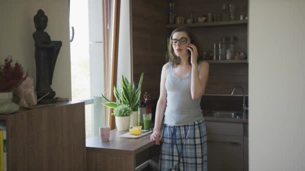 Молодая женщина разговаривает по смартфону стоя на кухне — стоковое видео