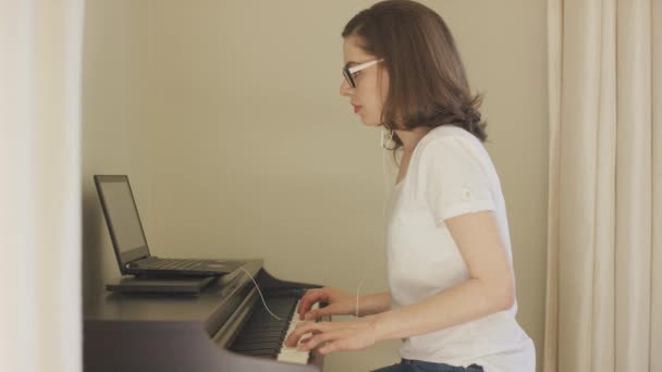 Молодая женщина играет на пианино с помощью ноутбука и наушников — стоковое видео