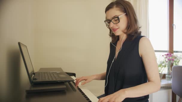 Веселая женщина играет на пианино с помощью ноутбука и наушников — стоковое видео