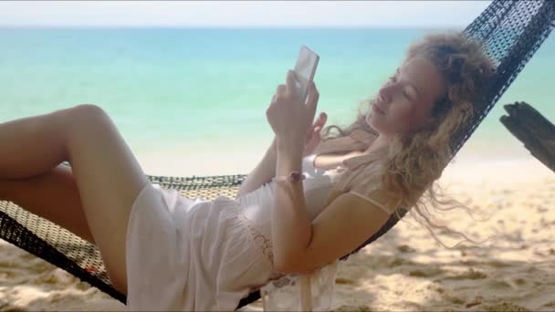 Entspannte Frau surft Smartphone, während sie in der Hängematte am Strand faulenzt — Stockvideo