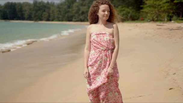 Mujer agradable sosteniendo vestido florecido y caminando en la playa de arena en día brillante — Vídeos de Stock