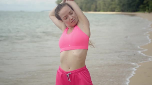 Femme concentrée en vêtements de sport roses étirant les mains levées sur la plage — Video