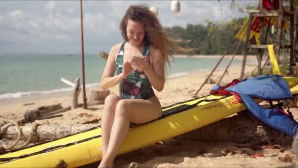 Улыбающаяся женщина в купальнике сидит на пляже и пользуется смартфоном — стоковое видео