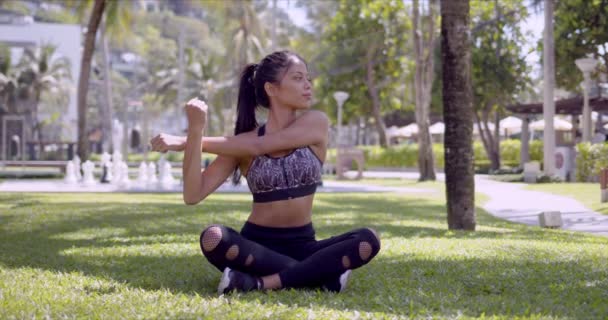 Asiática esportiva mulher sentado com pernas cruzadas e alongamento braços no parque — Vídeo de Stock