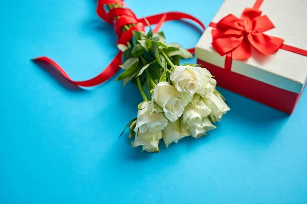 白色玫瑰的花束与红色弓在蓝色背景。侧面的盒装礼物 — 图库照片