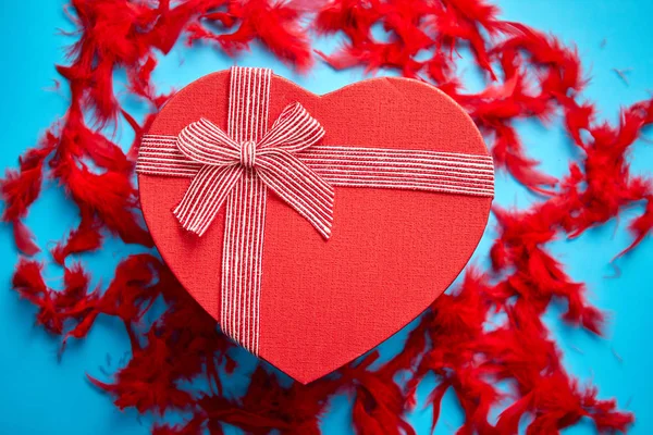 红色, 心形礼品盒放置在红色羽毛之间的蓝色背景 — 图库照片