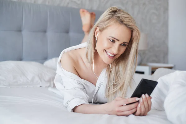 Adorable, mujer rubia sonriente acostada en la cama blanca y usando un teléfono inteligente —  Fotos de Stock