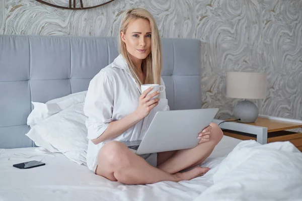 Glückliche schöne Frau arbeitet an einem Laptop sitzt auf dem Bett im Haus. — Stockfoto