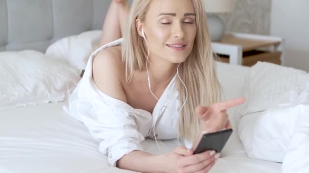 Очаровательная, улыбающаяся блондинка лежит в белой кровати и пользуется смартфоном. — стоковое видео