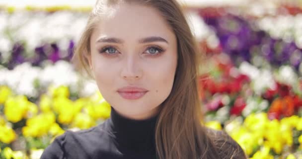 Vacker kvinna med blå ögon och makeup poserar på suddig bakgrund av färgglada blommor — Stockvideo