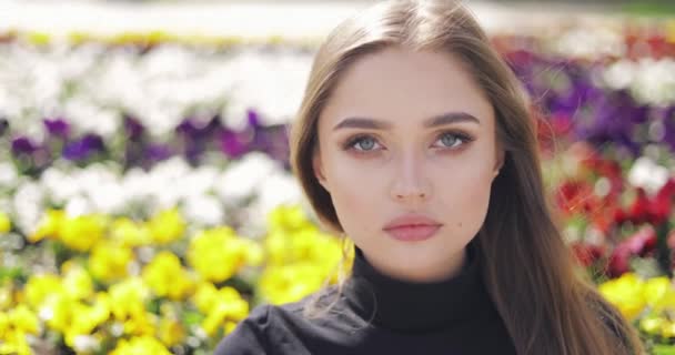 Mooie vrouw met blauwe ogen en make-up poseren op wazig achtergrond van kleurrijke bloemen — Stockvideo