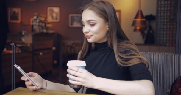 예쁜 소녀 가 카페에 앉아 손에 커피를 들고 스마트폰을 하고 있습니다. — 비디오