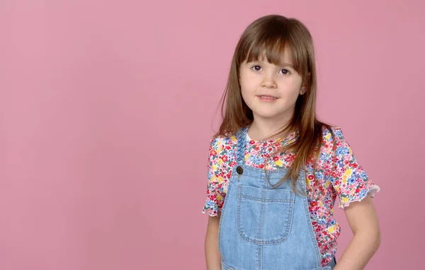 Söt liten flicka 6-7 år gammal poserar i fängelsehålor jeans och blomma mönster blus på rosa — Stockfoto