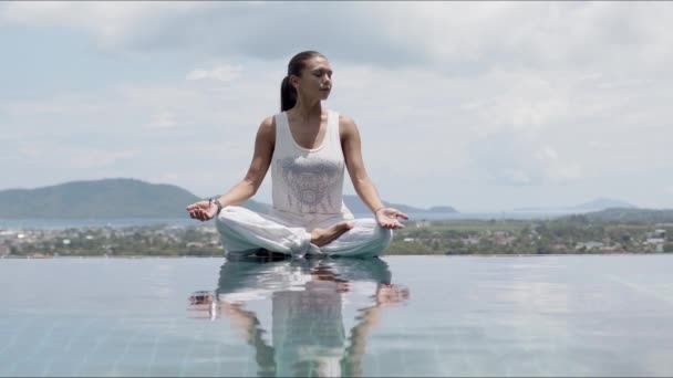 Femme sereine pratiquant le yoga en position lotus au bord de la piscine contre le ciel — Video