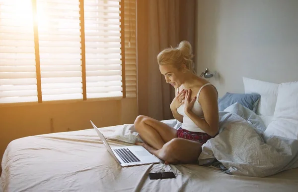 Encantadora mujer en pijama sentada en la cama cerca de la computadora portátil y mirándola —  Fotos de Stock