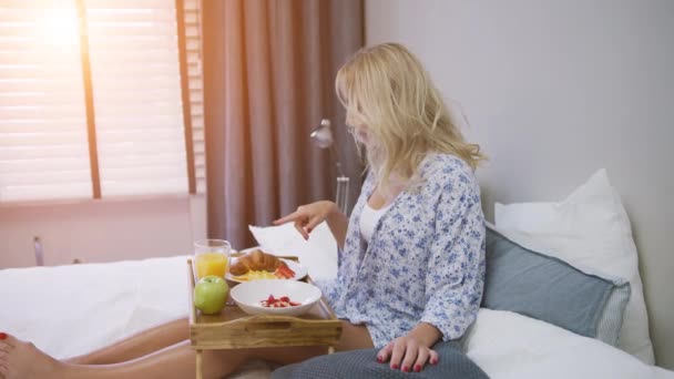 Vonzó fiatal nő ingben reggelizik, miközben az ágyon ül tálcával — Stock videók
