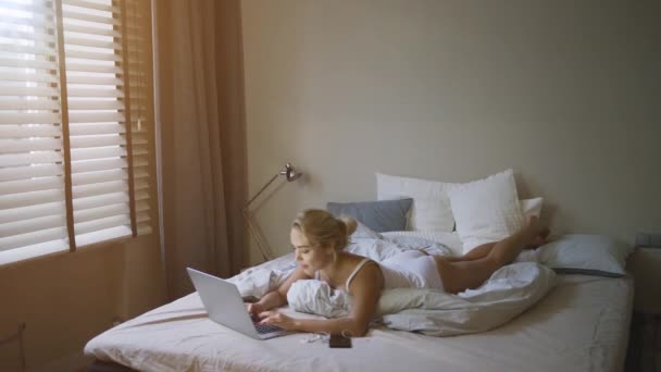 Sinnliche junge Frau in weißem Tank-Top und Höschen auf dem Bett neben Laptop liegend — Stockvideo