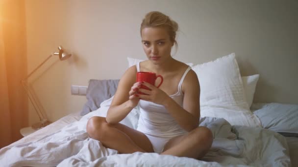 Mooie vrouw in wit tank top en slipje zitten op bed en genieten van warm drankje — Stockvideo