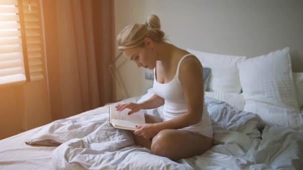Милая молодая женщина в белой майке и трусиках сидит на удобной кровати и читает — стоковое видео