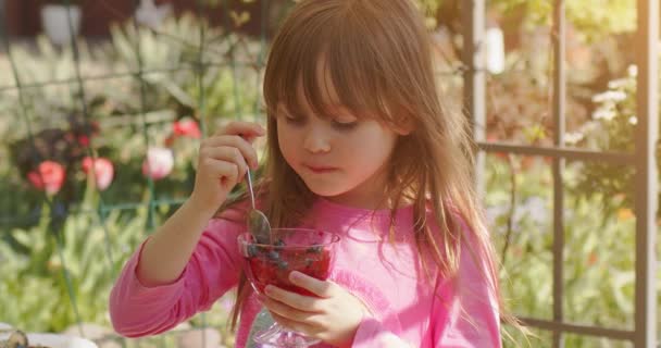 Söt liten 6 eller 7 år gammal flicka äter frukt dessert gelé i sommarträdgården. Långsam rörelse video — Stockvideo