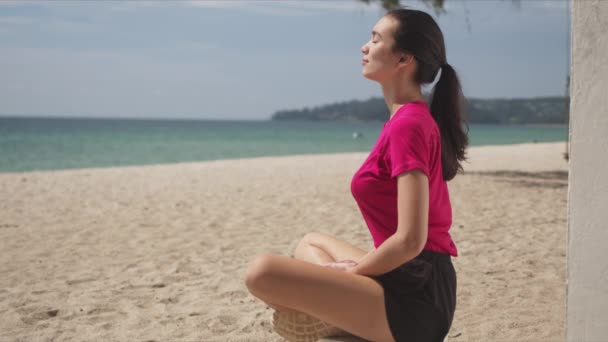 Vista lateral da jovem mulher esportiva vestindo camisa rosa e shorts pretos sentados na posição de lótus — Vídeo de Stock