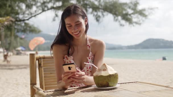 Młoda ciemnowłosa kobieta w letniej sukience sprawdza wiadomość na smartfonie siedząc w kawiarni na plaży — Wideo stockowe