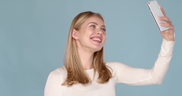 Retrato de uma jovem alegre com sorriso adorável tomando selfie isolado sobre fundo azul — Vídeo de Stock