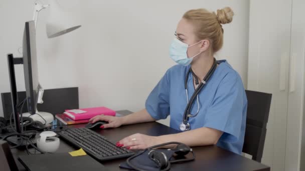 Впевнена досвідчена молода лікарка з маскою обличчя, що сидить перед комп'ютером в офісі — стокове відео