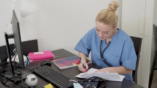 Молода красива блондинка-лікарка в синьому пальто, працює за столом за допомогою комп'ютера і робить паперові роботи — стокове відео