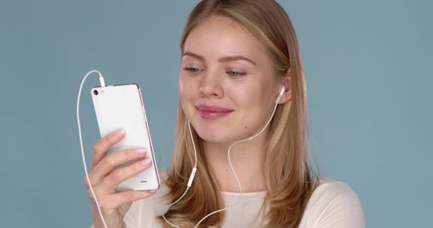 Mujer hermosa feliz posando aislada sobre la pared azul escuchando música con auriculares usando el teléfono — Vídeos de Stock