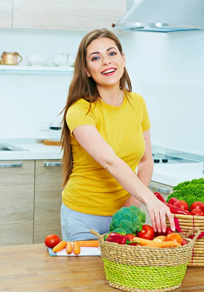 キッチンのかごに野菜をテーブルの近くに立っているカジュアルな服を着て長髪の若い女性を笑顔 — ストック写真
