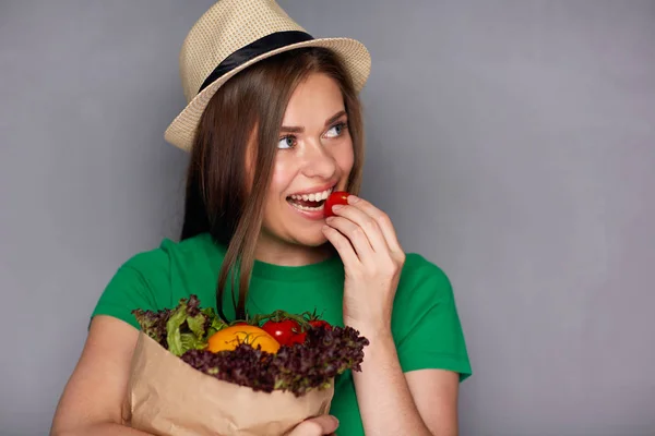 野菜が入った紙袋を押しながら灰色の壁の背景にトマトを食べる若い女性 — ストック写真