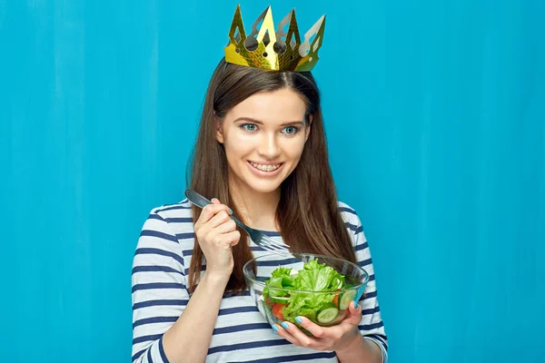 smiling woman in crown holding bowl with green salad on blue wall background