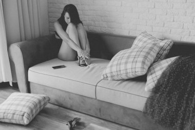 Makyajsız bir kadın kanepede oturuyor ve ruh hali hakkında kendini eleştiriyor. Olumsuz düşünceleri olan depresif bir atış. Efekt olarak tahıl eklenmiş siyah beyaz resim.