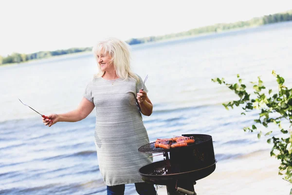 Kadın Kıdemli Izgara Sosis Göl Yakınındaki Yaz Mutfağı Yemek Hazırlama — Stok fotoğraf