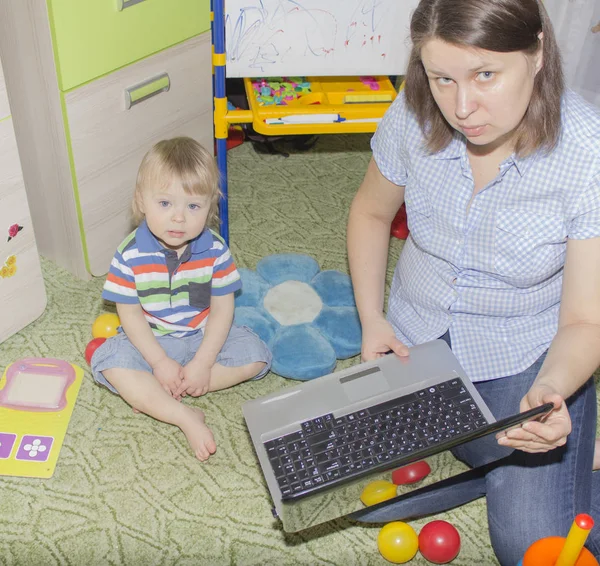 Lindo niño divertido bebé con pelo largo y rubio rizado jugando en la computadora y el teléfono móvil —  Fotos de Stock